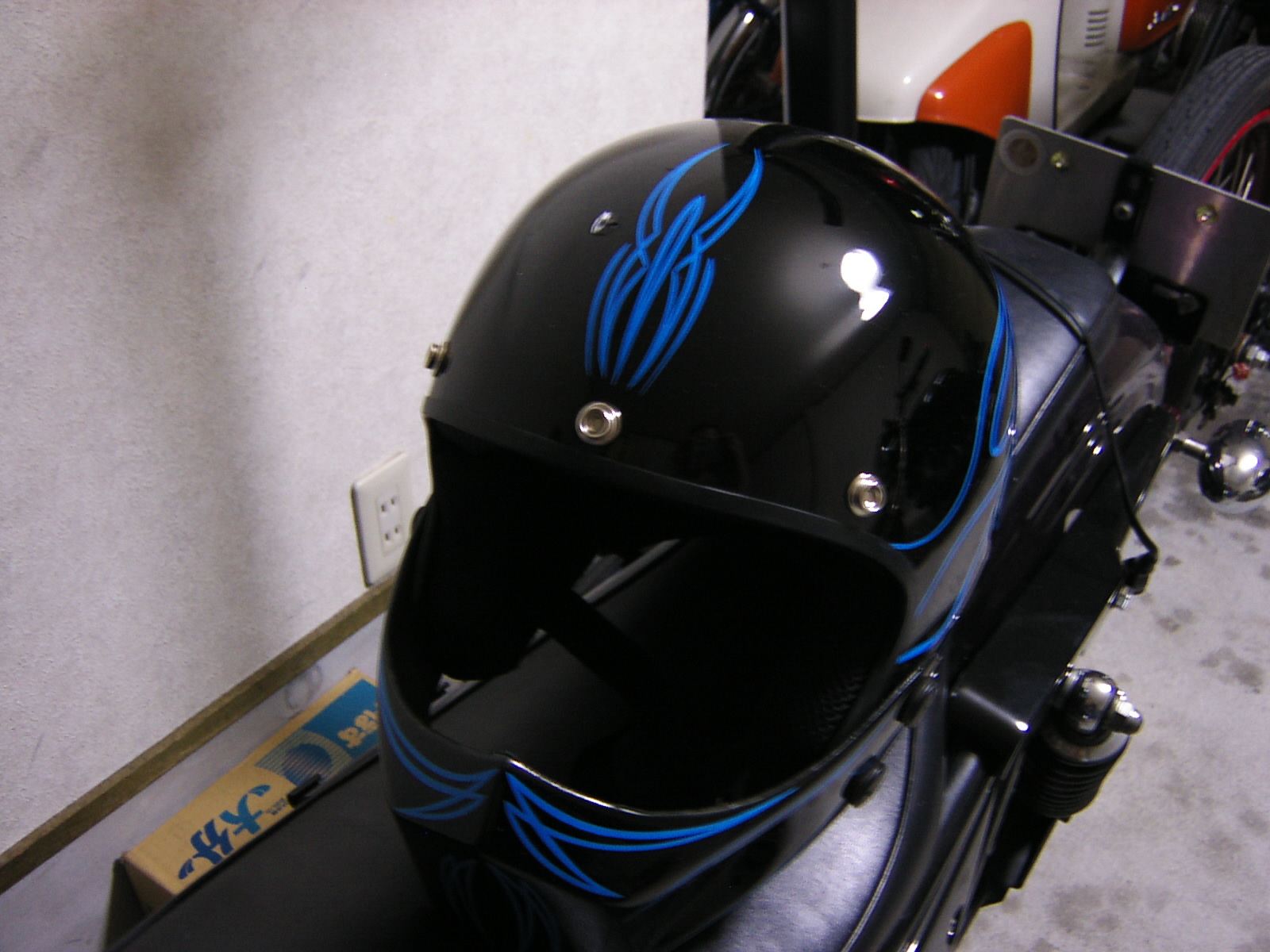 Black motorcycle helmet with blue pinstripe flame design, photographed indoors near motorcycle parts and a wall outlet
